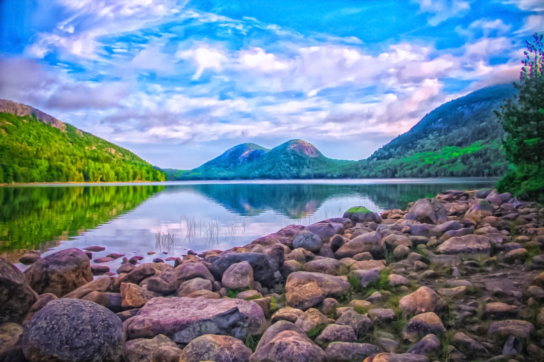 Rocky shoreline along a calm lake with forested mountains in the background, under a partly cloudy sky. The water reflects the sky and surrounding greenery, creating a peaceful, natural landscape scene.