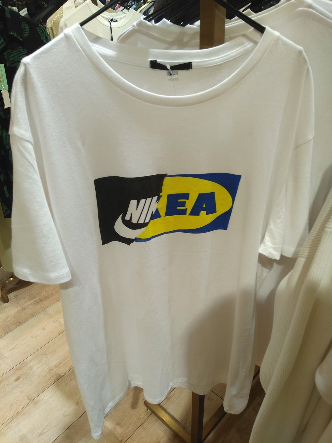 A white T-shirt hangs on a rack, featuring a logo that combines the Nike "swoosh" and text with the blue and yellow IKEA logo design.