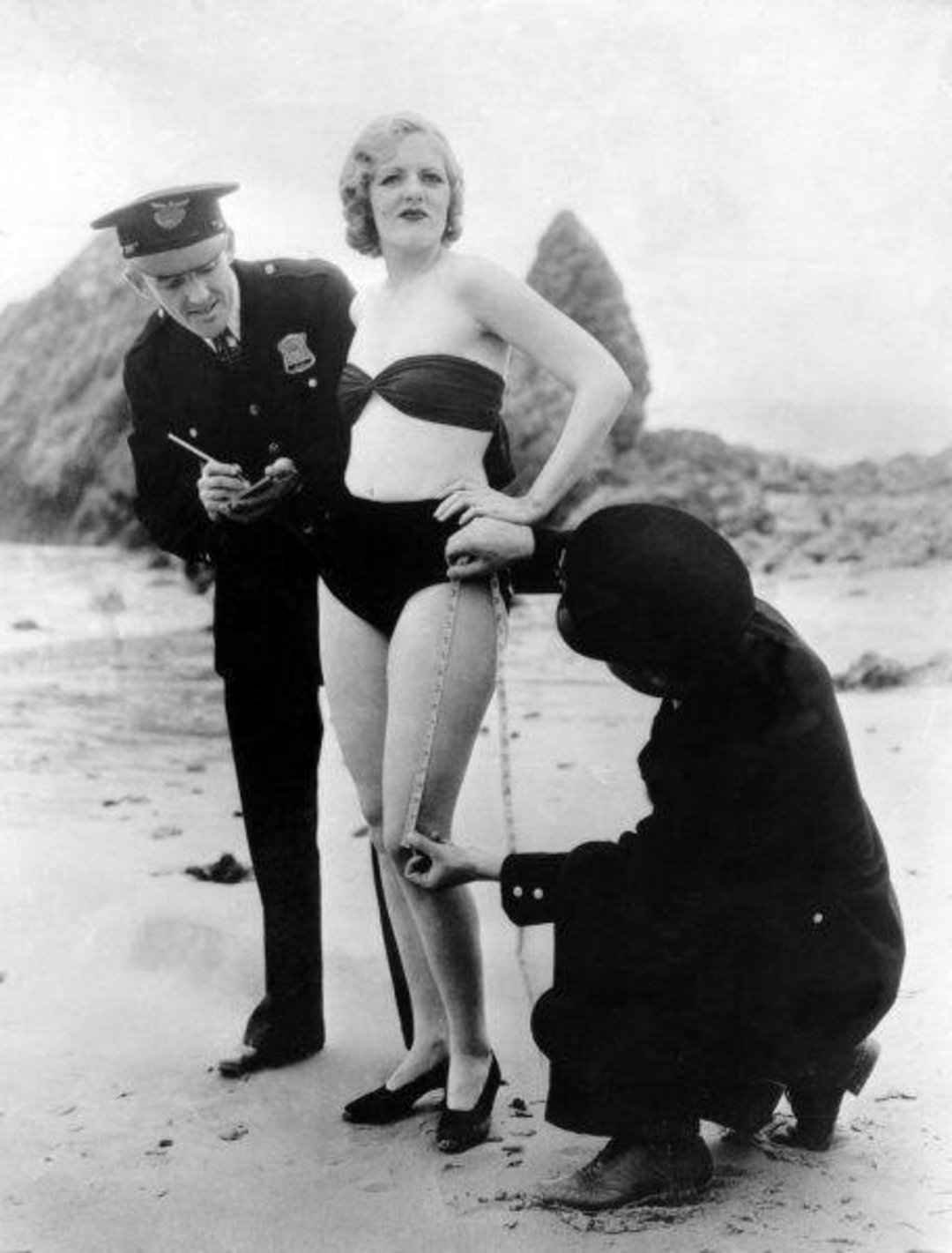 A woman in a swimsuit stands on a beach while a police officer takes notes and another person measures her thigh with a tape measure.
