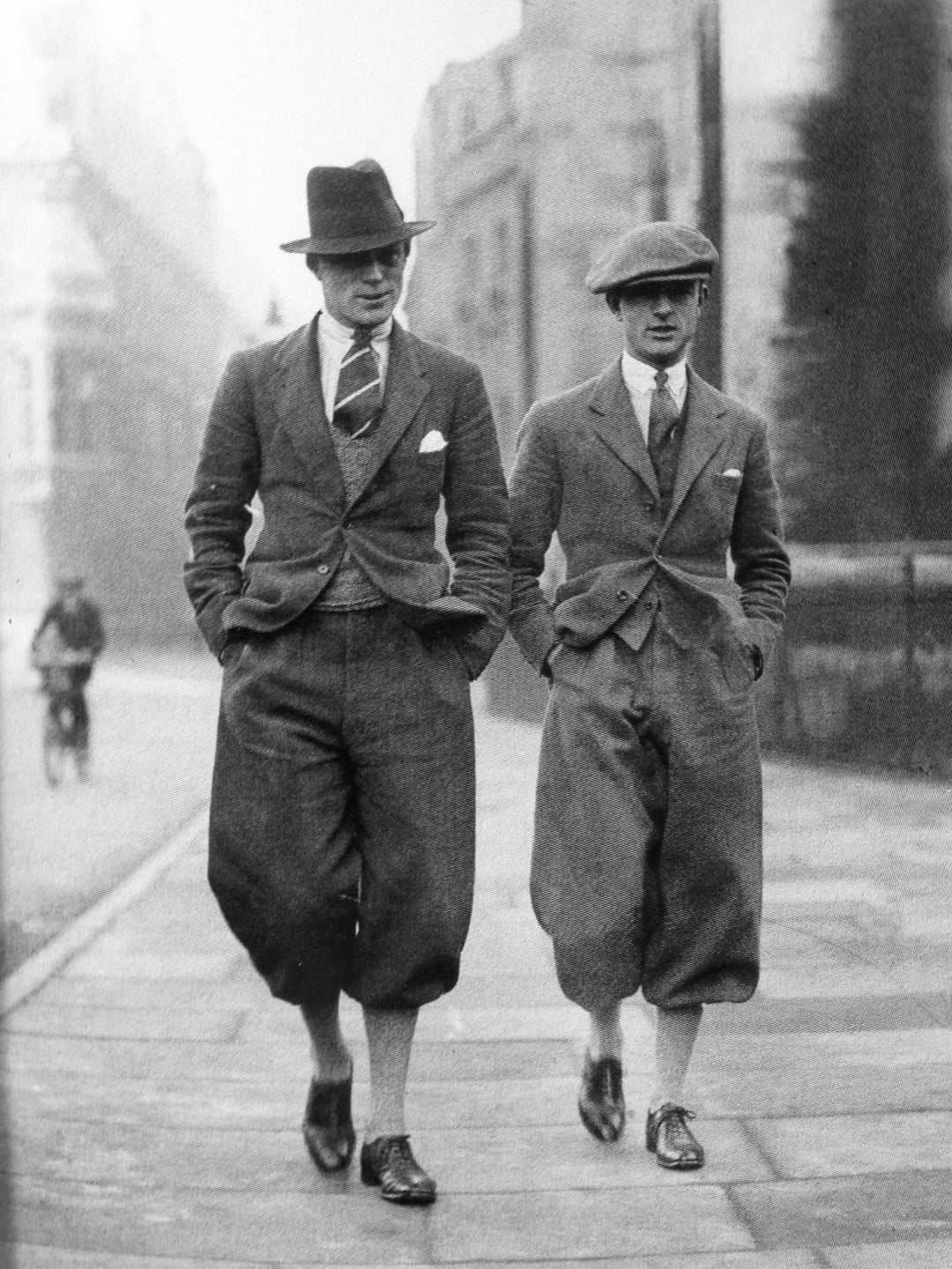 Two men in baggy suits, ties, and hats walk side by side on a city sidewalk in the early 20th century. Both have hands in pockets, and a cyclist is visible in the background.