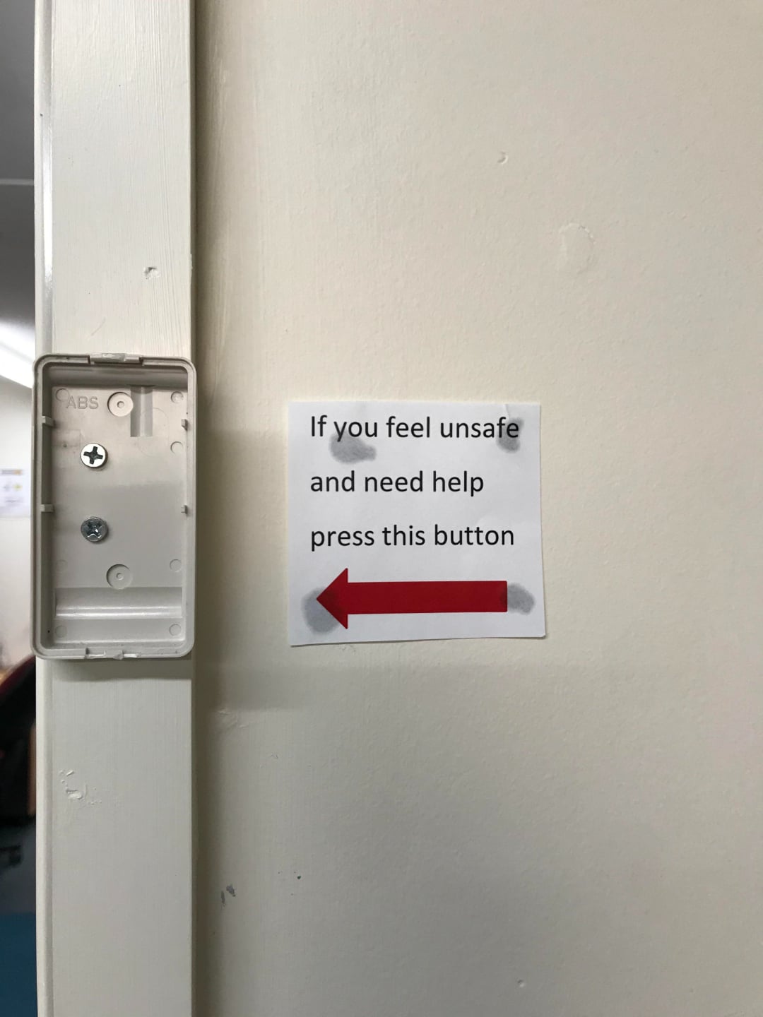 A sign on a wall reads, "If you feel unsafe and need help press this button," with a red arrow pointing left to an empty box where a button is missing.
