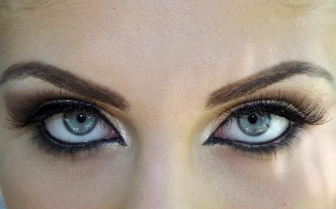 Close-up of a person's face, focusing on their blue eyes, dramatic dark eyeliner, long eyelashes, and well-defined eyebrows. The makeup creates a bold, intense look.