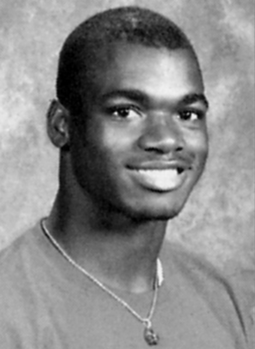 Black and white portrait of a young man smiling at the camera. He has short hair and wears a necklace and a plain shirt, posed against a mottled studio background.