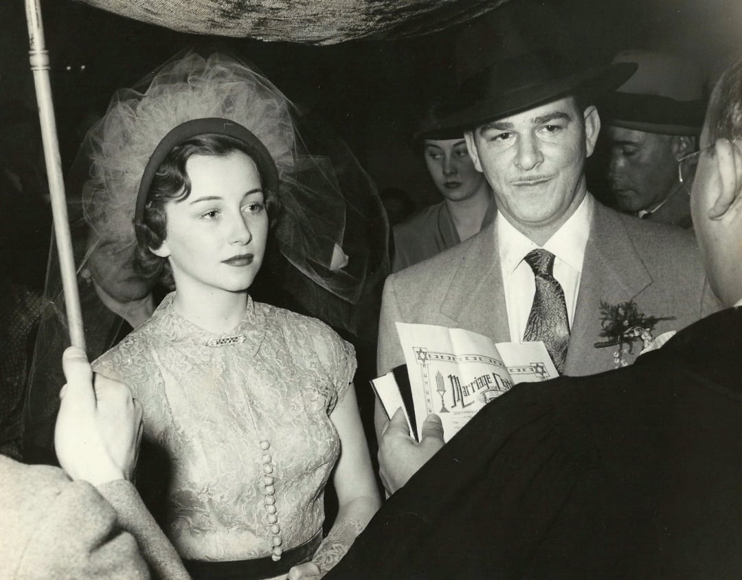 A bride and groom stand together under an umbrella during a wedding ceremony. The bride wears a veil and lace dress, while the groom is in a suit and hat. People surround them, and someone holds an open book in front of the couple.