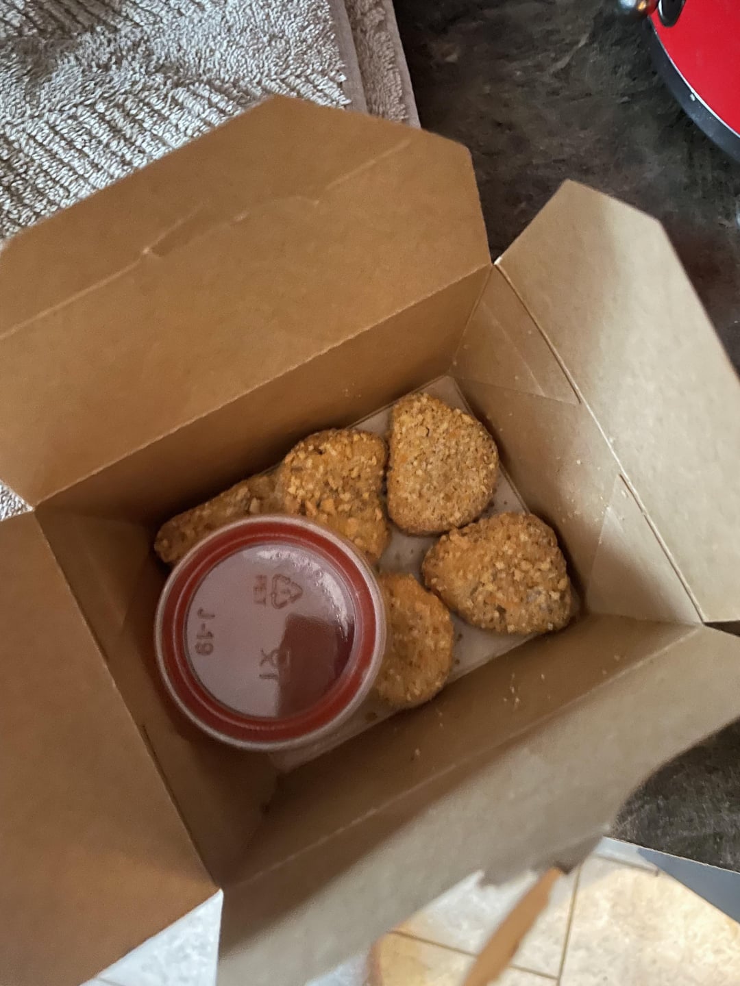 A brown takeout box containing six breaded chicken nuggets and a small cup of dipping sauce, placed on a kitchen counter near a towel.