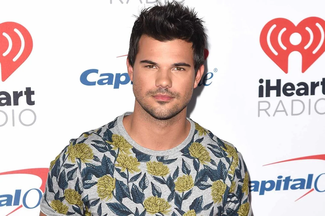 A man with short dark hair and facial hair stands in front of an iHeartRadio event backdrop, wearing a grey shirt with a yellow floral pattern.