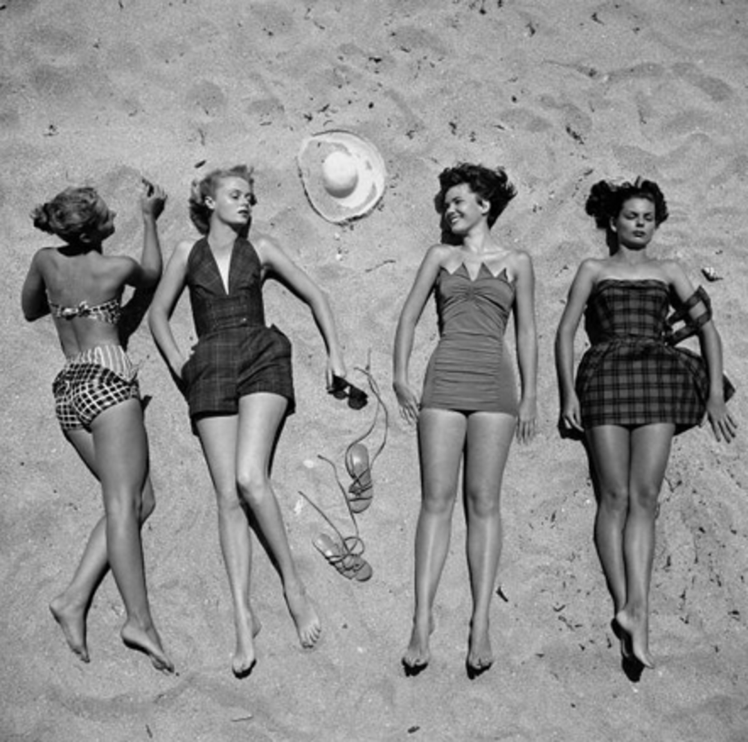 Four women in vintage swimsuits lie on sandy beach, side by side, with a hat, a pair of sunglasses, and sandals on the sand beside them. They appear relaxed and are facing upward, enjoying the sun.