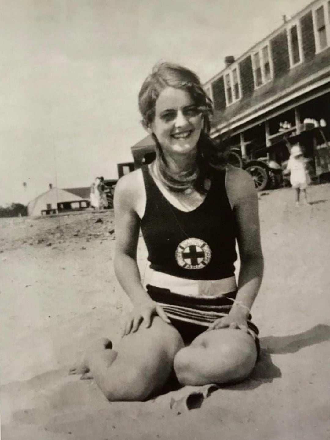 A young woman in a vintage swimsuit kneels and smiles on a sandy beach. Behind her are old-fashioned buildings, a car, and people in period clothing, suggesting the photo is from the early 20th century.