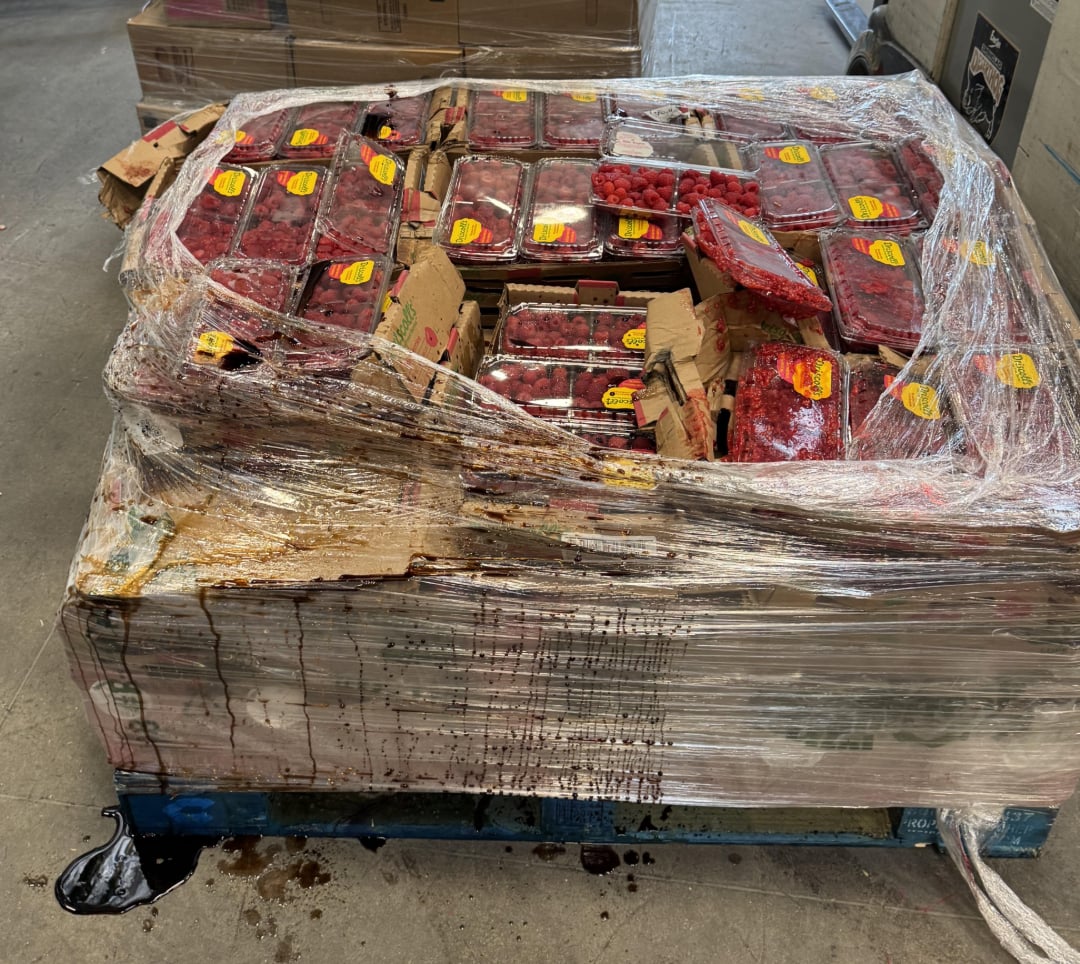 A pallet wrapped in plastic holds multiple damaged boxes of raspberries, with many containers crushed and leaking juice, which pools and drips onto the warehouse floor.