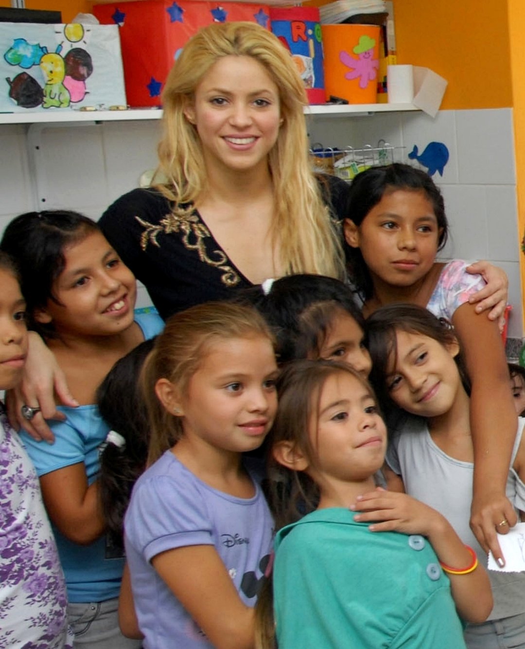 A woman with long blonde hair stands smiling and hugging a group of young girls, all smiling, in a colorful indoor setting decorated with children's artwork.