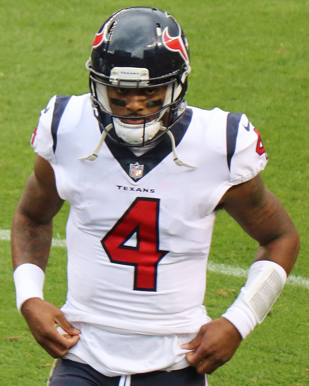 A football player in a white Houston Texans uniform with the number 4 on the front stands on a field, wearing a dark helmet and gloves, with a taped wrist and a determined expression.