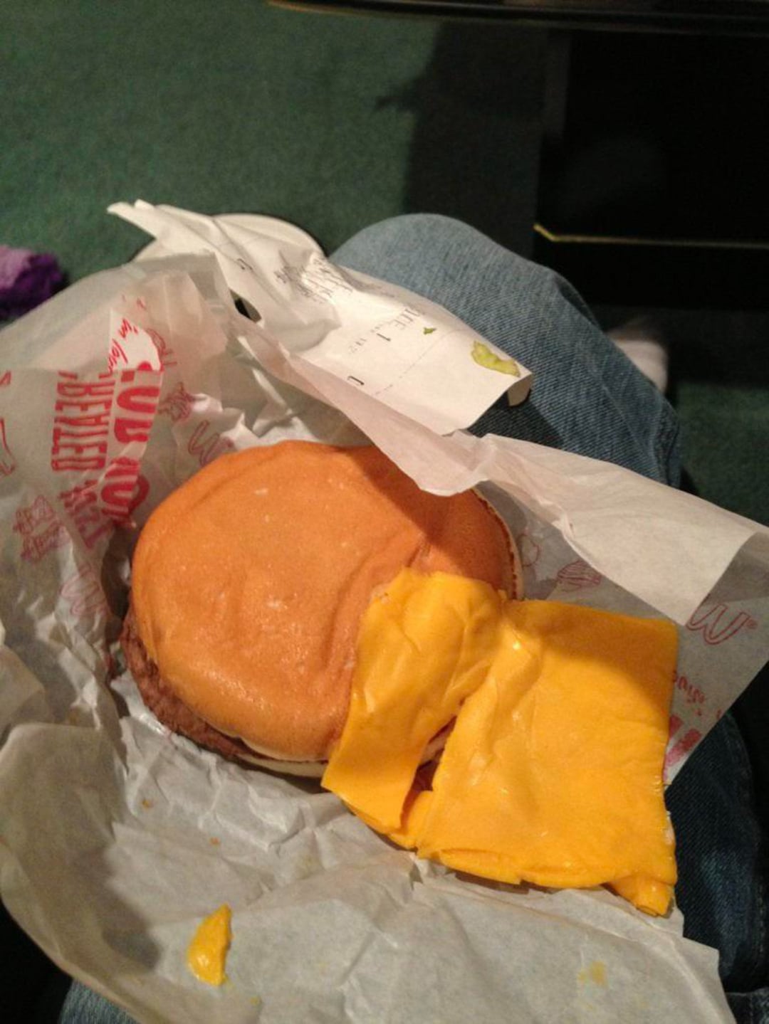 A cheeseburger with the cheese slice mostly stuck to the wrapper sits on someone's lap, leaving the burger bun and patty mostly bare.