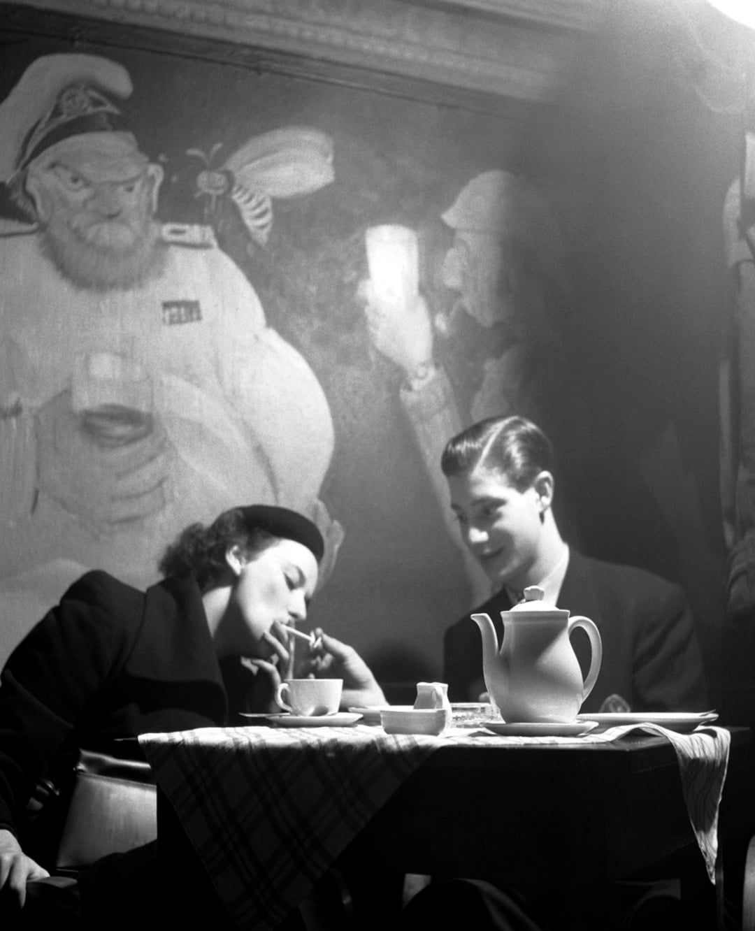 A woman and man sit at a table with a teapot and cups, sharing tea and pastries. The woman smokes a cigarette. A mural of a bearded sailor and another figure is visible on the wall behind them.