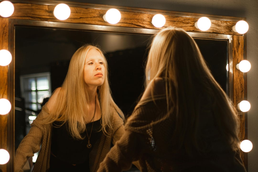 A woman with long blonde hair looks at her reflection in a mirror framed by round vanity lights, wearing a black top and a beige sweater, in a softly lit room.