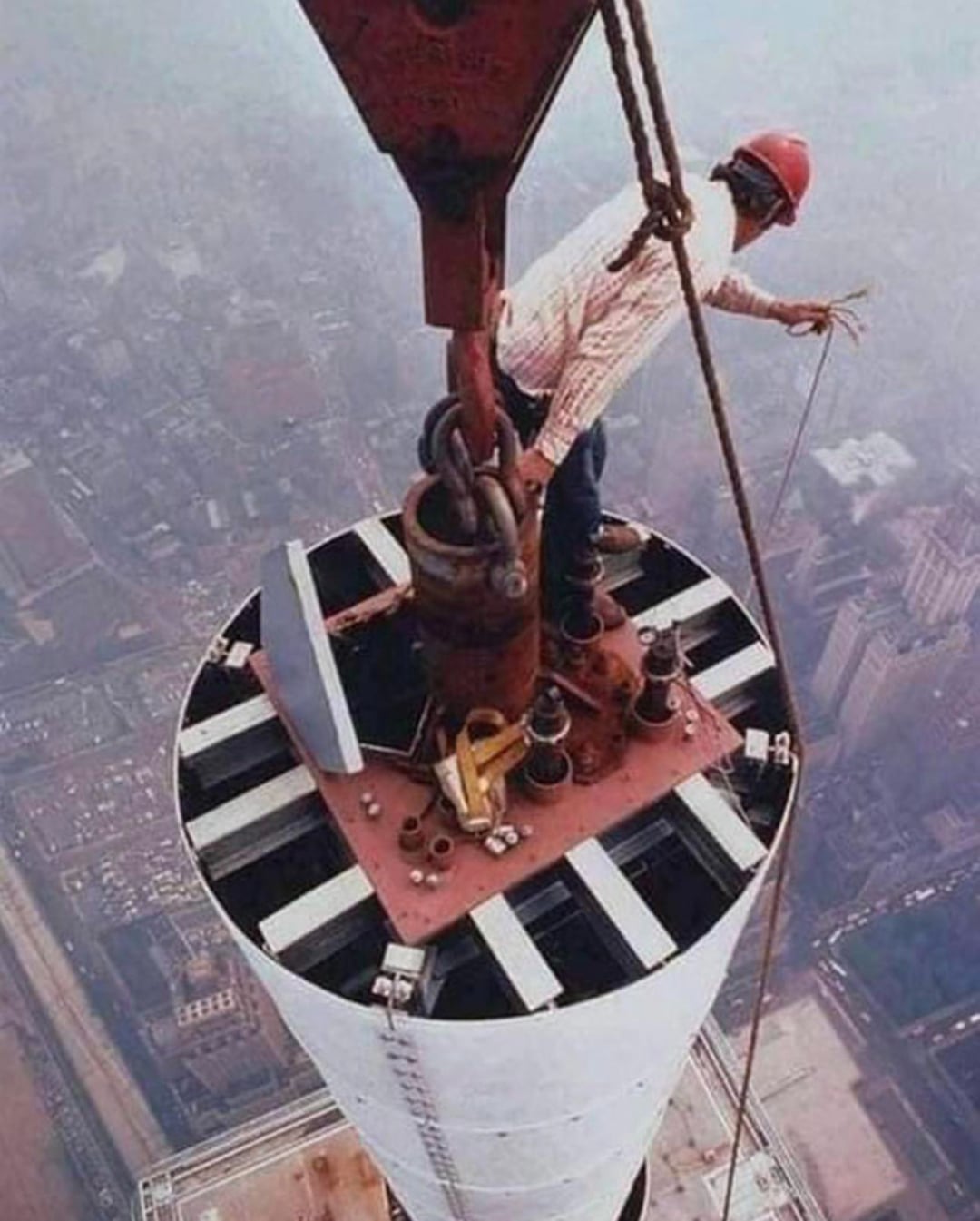 A construction worker in a red hard hat stands on top of a tall structure, holding onto a safety rope, high above a city, with buildings and streets visible far below through the haze.