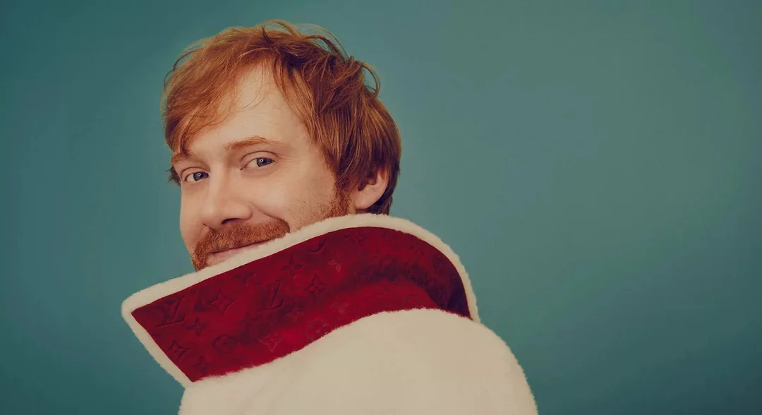 A man with light red hair and a beard smiles over his shoulder, wearing a white coat with a high red collar, against a plain teal background.
