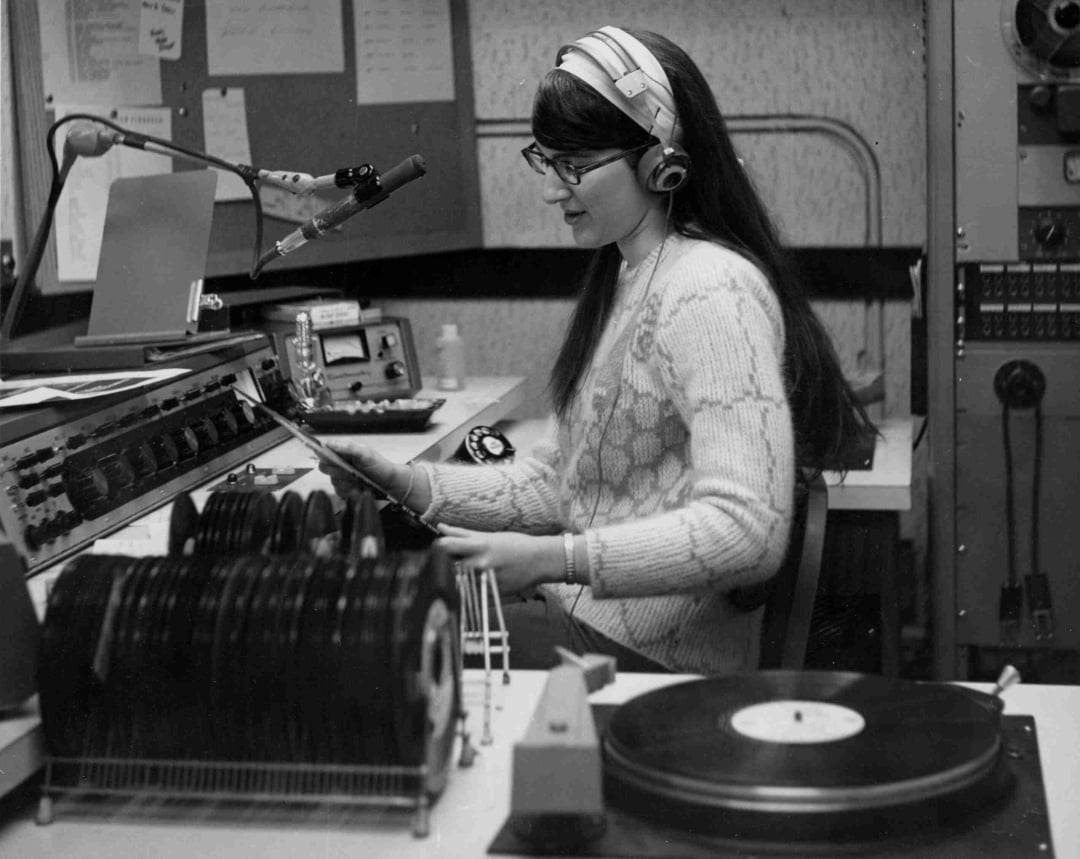 A woman with long hair and glasses sits at a radio station control panel, wearing headphones and holding a vinyl record. She is surrounded by audio equipment, a microphone, and stacks of records.