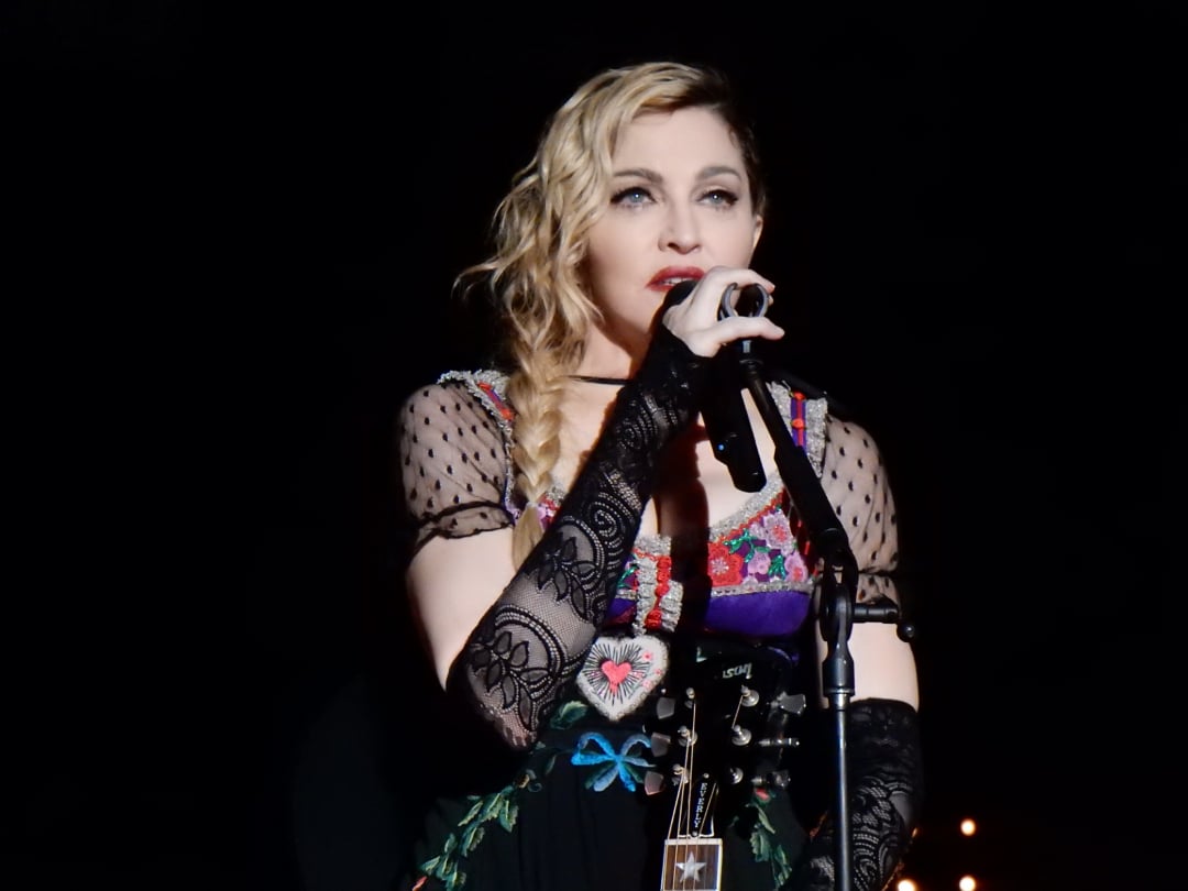 A woman with wavy blonde hair and lace gloves sings into a microphone on stage, wearing a floral embroidered dress with sheer polka dot sleeves. A guitar hangs from her shoulder, and the background is dark.