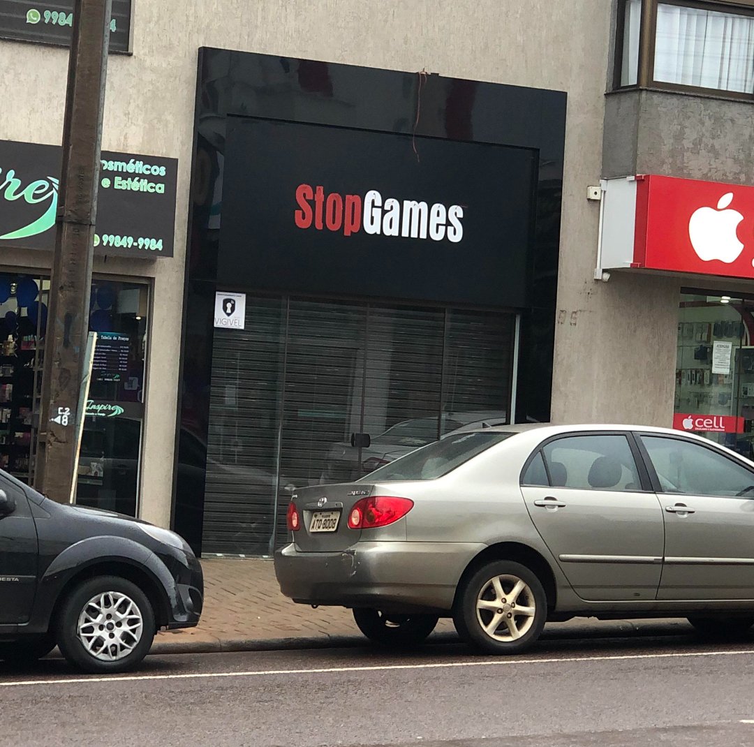 A storefront with a black sign that reads "StopGames" in red and white text. Two cars are parked on the street in front, and neighboring shops are partially visible.