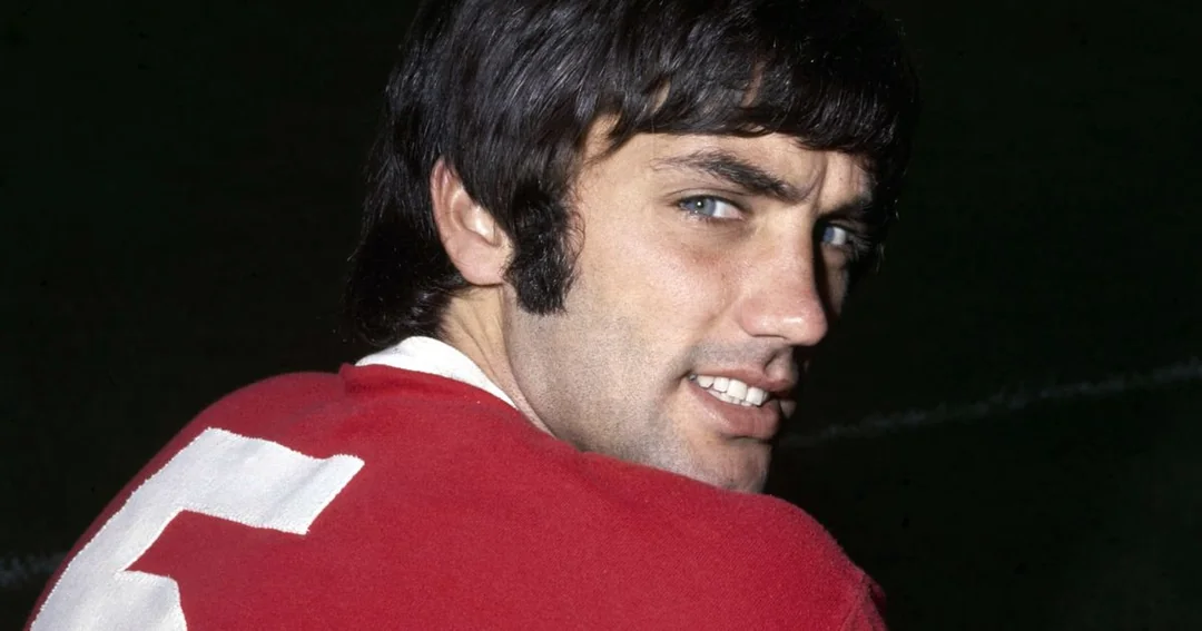 A man with dark hair and sideburns, wearing a red sports jersey with a white number 7 on the back, looks over his shoulder and smiles slightly against a dark background.