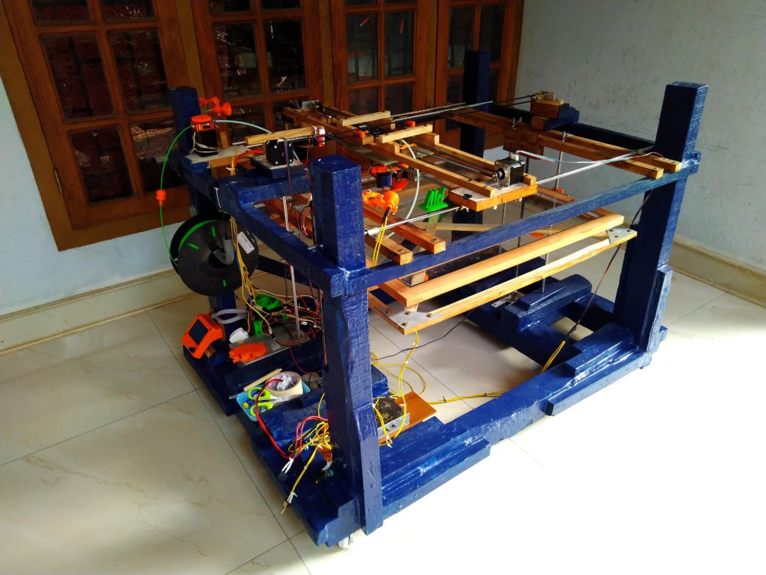A homemade 3D printer with a blue wooden frame, exposed wires, electronic components, and various mechanical parts sits on a tiled floor near a wooden window inside a room.