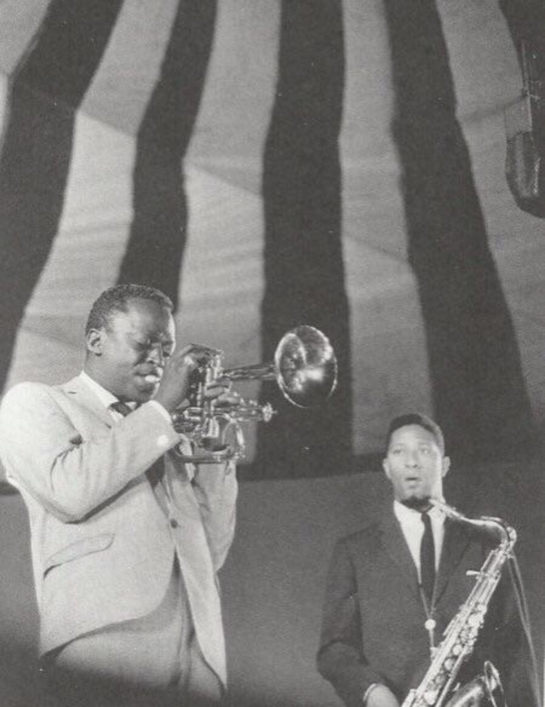 Two jazz musicians in suits perform on stage; one plays the trumpet with eyes closed, while the other stands next to him holding a saxophone. Large striped patterns are visible on the ceiling above them.