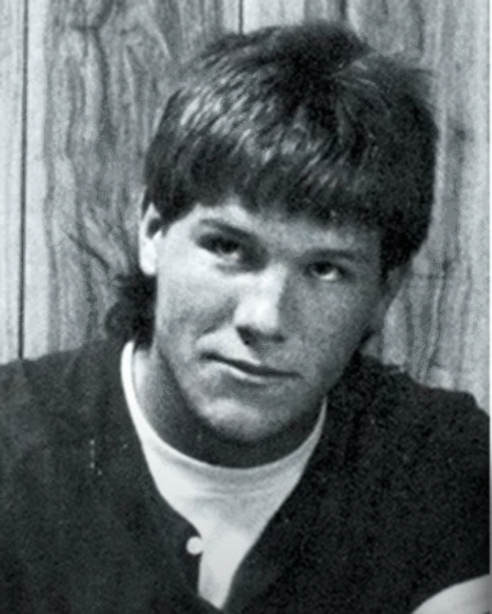 A young person with short, slightly messy hair looks at the camera, wearing a dark shirt over a light undershirt, posed in front of a wood-paneled wall. The image is in black and white.