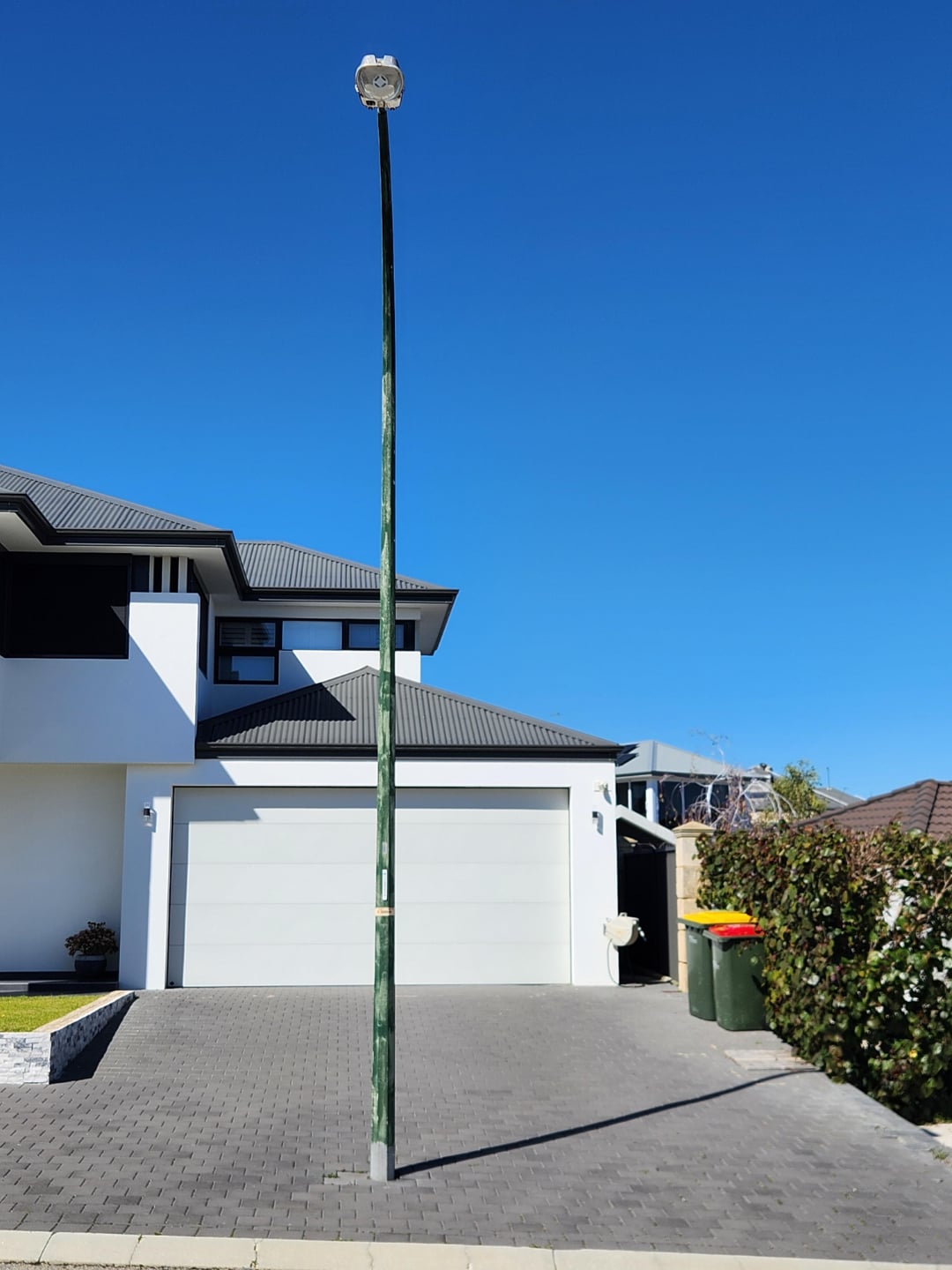 A green streetlight pole is bent and leaning forward in the middle of a driveway in front of a modern white house with a double garage, under a clear blue sky.