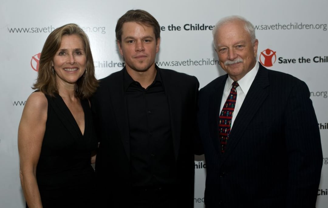 Three people stand together dressed in formal attire, posing for a photo in front of a "Save the Children" step-and-repeat banner.