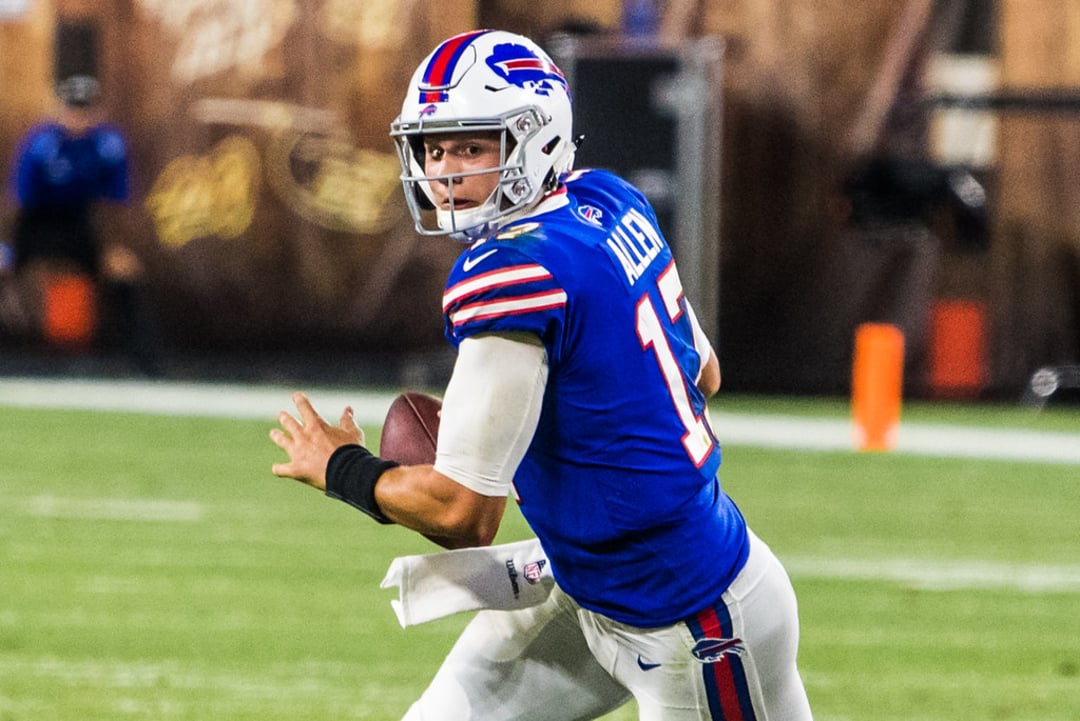 A football player in a blue Buffalo Bills uniform prepares to throw a pass during a game. He is running with the football, looking downfield, with a blurred stadium background behind him.