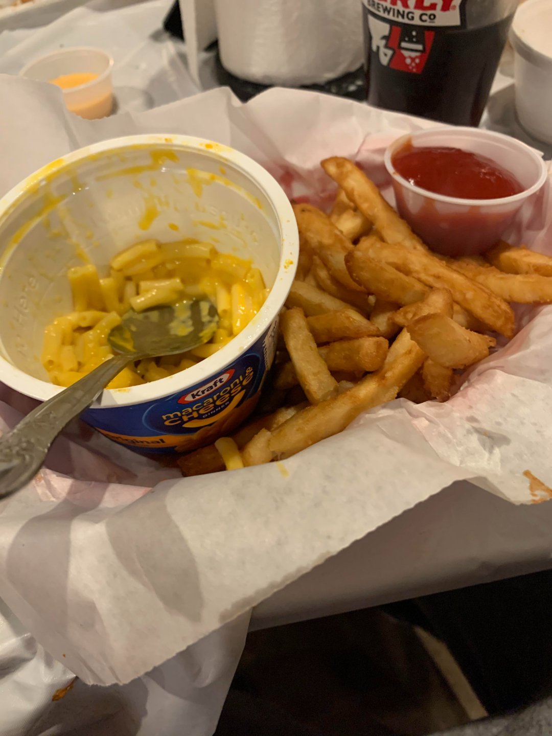 A tray of crinkle-cut French fries next to a cup of Kraft Easy Mac and cheese with a spoon inside, and a small container of ketchup on the side. The tray is lined with parchment paper.