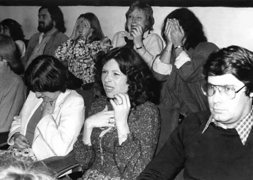 A group of people in a theater audience show mixed reactions—some appear amused, while others cover their faces or look anxious, suggesting they are watching an intense or suspenseful scene.