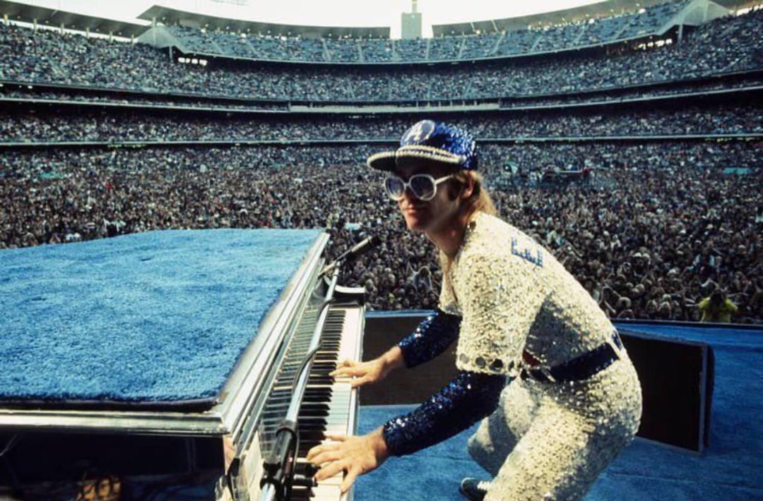 A performer in a glittery, sequined outfit and large sunglasses plays a grand piano on stage before a massive stadium crowd during an outdoor concert.