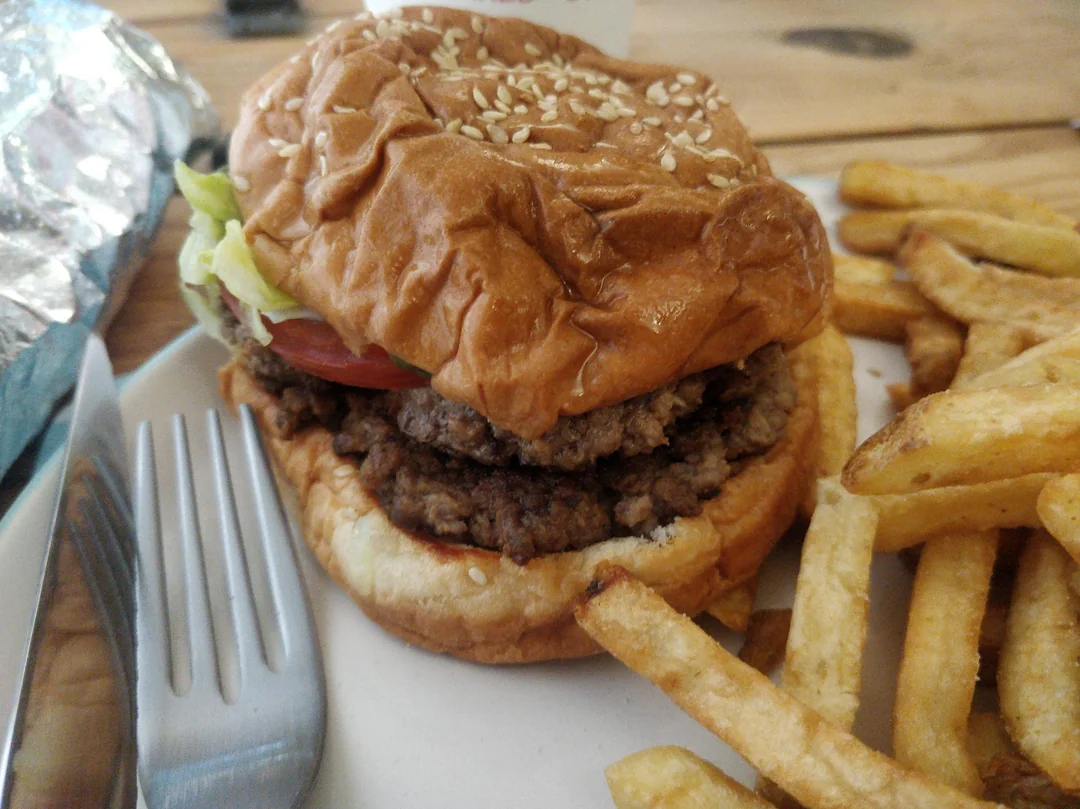 A double cheeseburger with lettuce and tomato in a sesame seed bun is served on a plate next to a portion of golden French fries. A fork rests on the plate beside the burger.