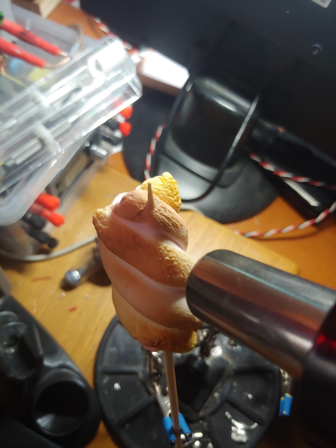 A close-up of a small swirl of meringue on a stick being toasted with a kitchen torch on a cluttered desk with tools and wires in the background.