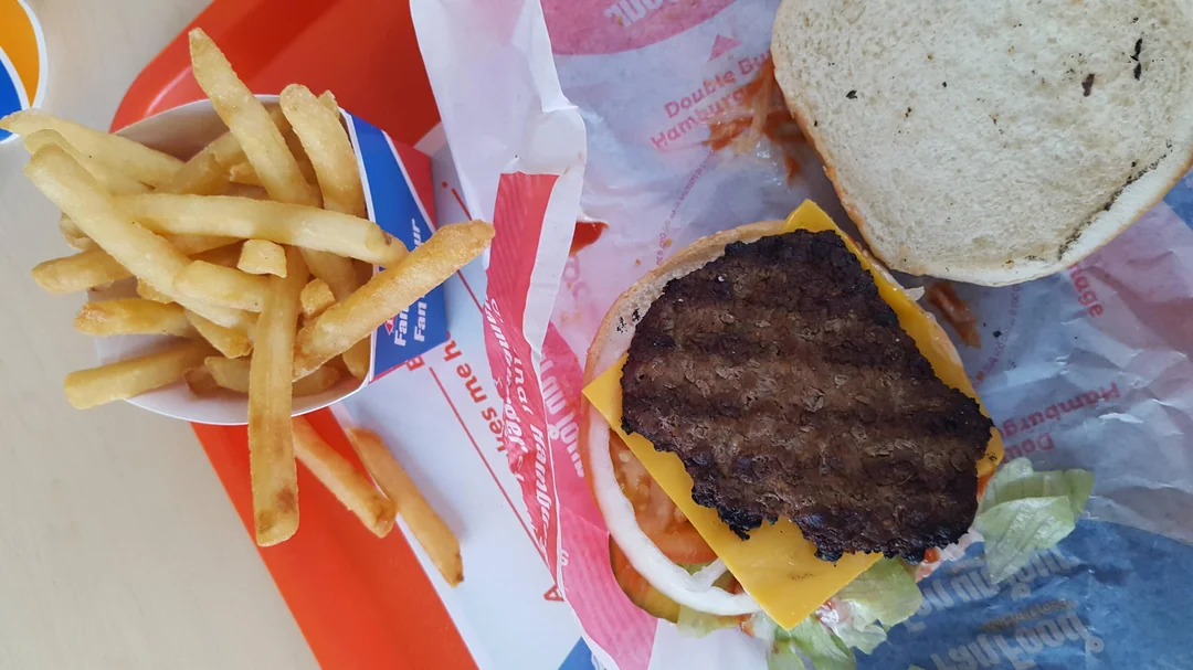 A tray with a cup of French fries and an open cheeseburger showing a beef patty, cheddar cheese, lettuce, tomato, and onion, with the top bun placed to the side.