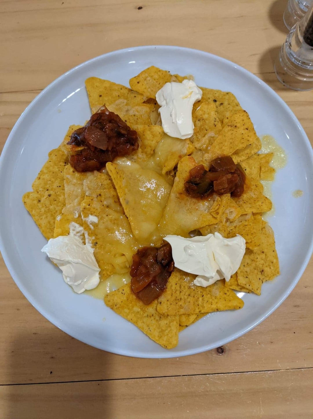 A plate of nachos topped with melted cheese, dollops of sour cream, and spoonfuls of chunky salsa sits on a wooden table.