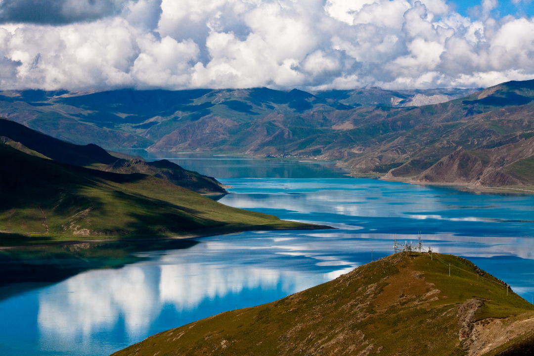 A vibrant blue lake winds through a mountainous landscape under a partly cloudy sky, with green hills and peaks reflected in the calm water.