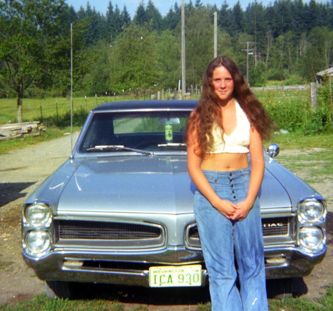 A young woman with long brown hair, wearing a white halter top and blue jeans, stands in front of a classic silver Pontiac car with a Washington license plate, in a rural outdoor setting with trees in the background.