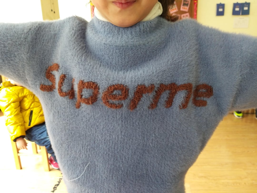 A child wearing a fuzzy light blue sweater with the word "Superme" in brown letters across the chest, standing indoors with arms outstretched.