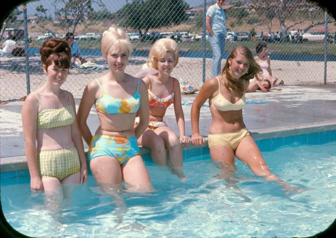 Four young women in colorful 1960s-style swimsuits sit at the edge of a pool, smiling. The background shows a chain-link fence, parked cars, trees, and people relaxing on the grass.