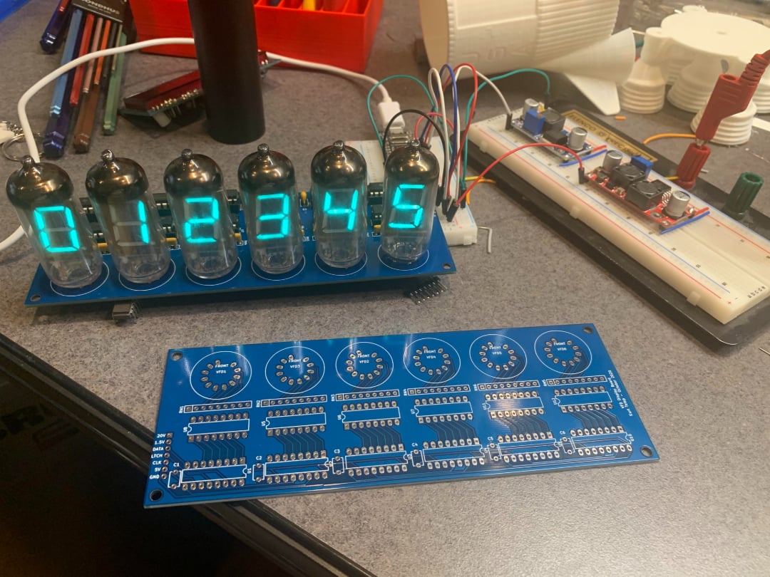 A nixie tube clock displaying digits 0 to 5 sits on a table next to a blue circuit board and a breadboard with wires and electronic components. Various tools and parts are visible in the background.