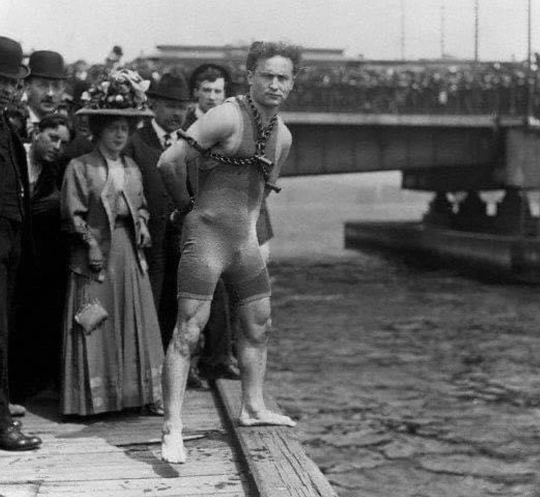 A man in a fitted suit, bound in chains, stands barefoot on a pier beside onlookers. A bridge and a crowd are visible in the background near the water.