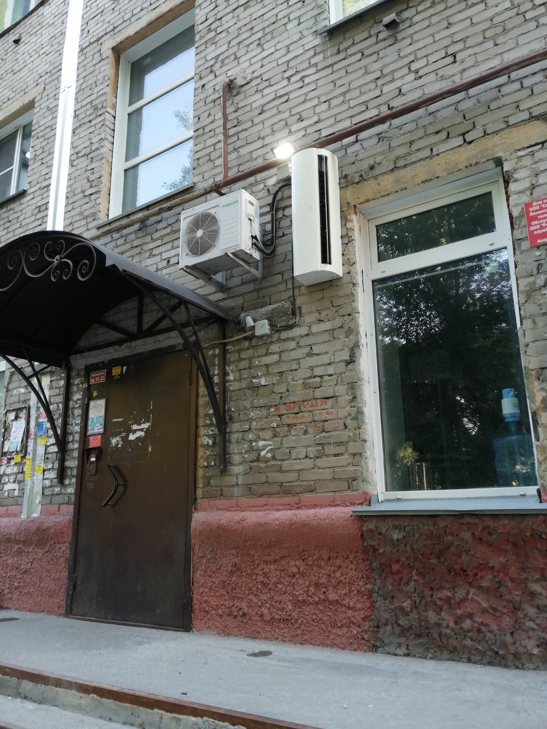 A metal door with a black awning leads into a brick building. An air conditioning unit is mounted above the door. A large window is to the right, reflecting trees. The lower wall is painted red.