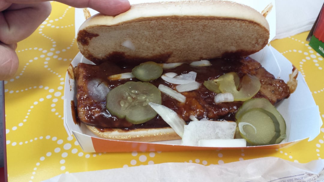 A hand lifts the top bun of a sandwich, revealing barbecue sauce-covered meat, pickle slices, and chopped onions inside a cardboard container on a yellow patterned surface.