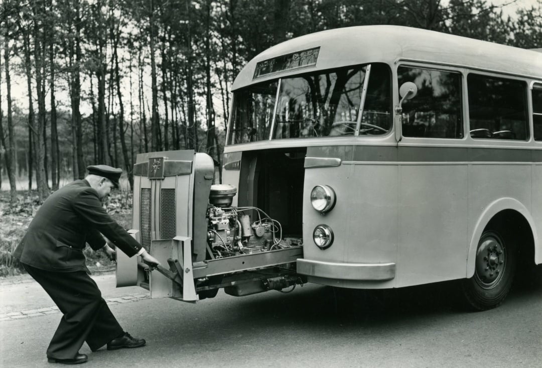 A man in a suit and hat pulls out the engine compartment from the front of a vintage bus, revealing its mechanical parts; trees and road are visible in the background.