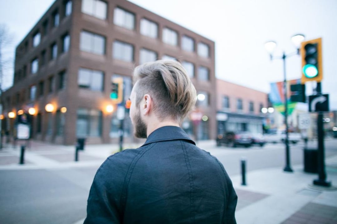A person with short, styled hair and a dark jacket walks along a city street near a brick building, streetlights, and traffic signals on a cloudy day. The photo is taken from behind.