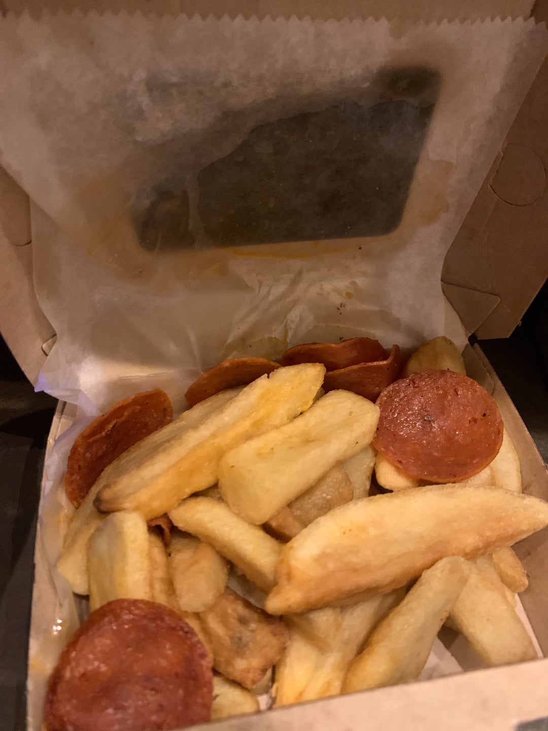 A cardboard takeout box with thick-cut fries and a few slices of pepperoni on top, lined with white parchment paper.