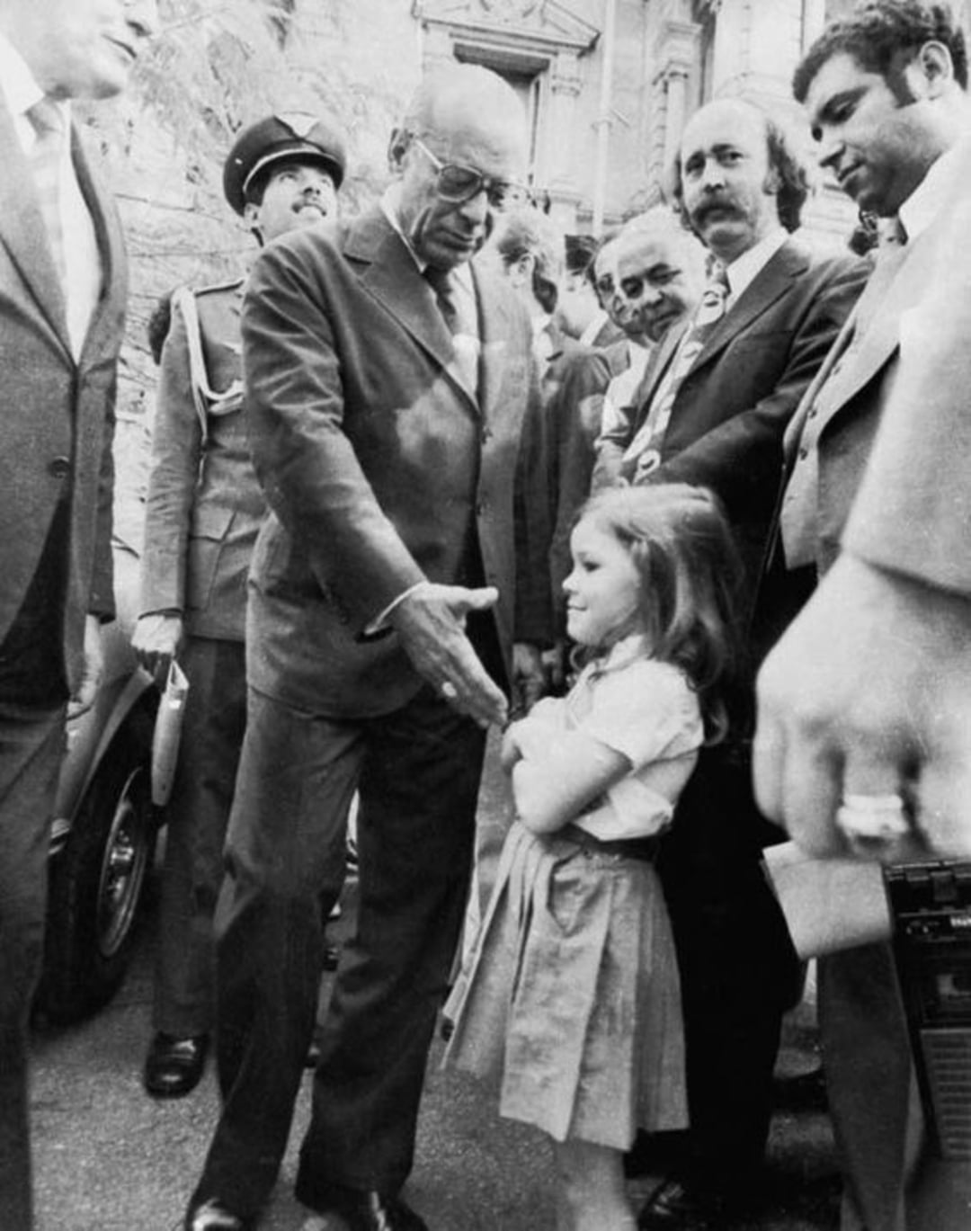 A man in a suit leans forward to shake hands with a young girl in a skirt, surrounded by smiling men in suits and a police officer in the background. The scene appears to be outdoors in front of a stone building.