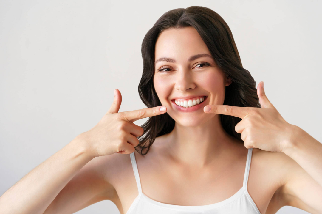 A smiling woman with long dark hair points to her teeth with both index fingers. She is wearing a white tank top and standing against a plain light background.